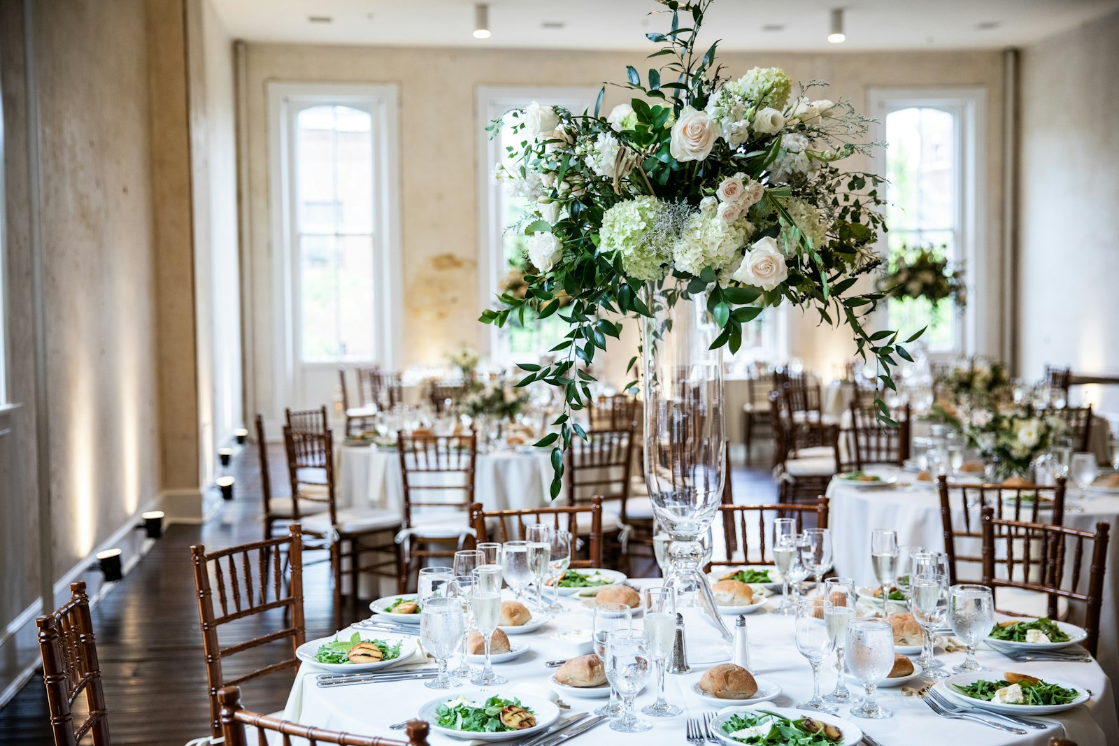A bright reception room with a tall white floral centrepiece and dressed round tables