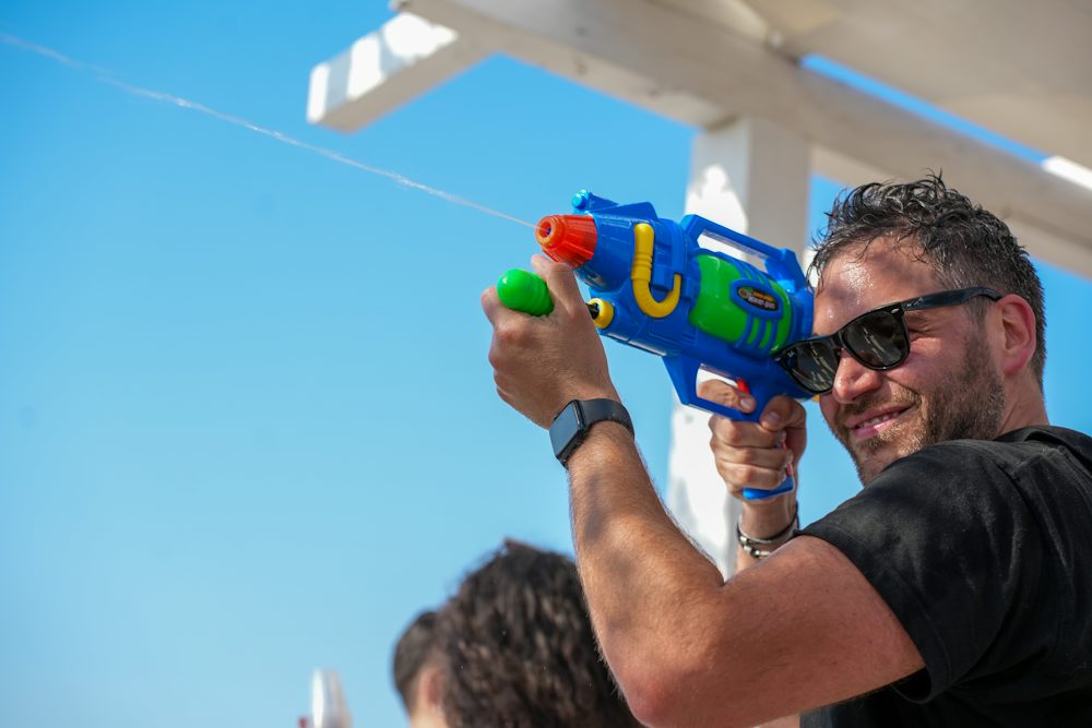 A smiling man firing a colourful water gun under a blue sky