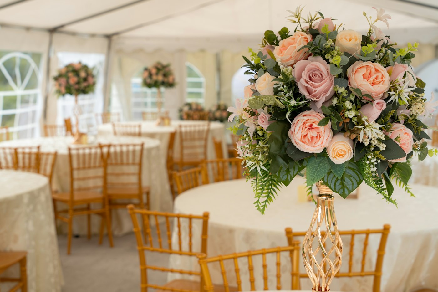 Round tables with gold chiavari chairs and rose centrepieces inside a white event tent